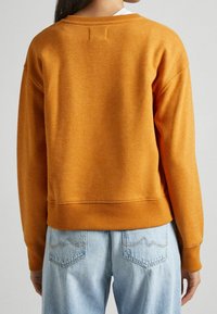 Person klädd i en senapsgul tröja med långa ärmar och ljusblå högmidjade jeans med bakfickor och subtila sömnadsdetaljer.