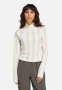 Biela športová mikina na zips s dlhými rukávmi, s jemnými vertikálnymi pruhmi a logom na hrudi, doplnená o šedé pruhované nohavice.