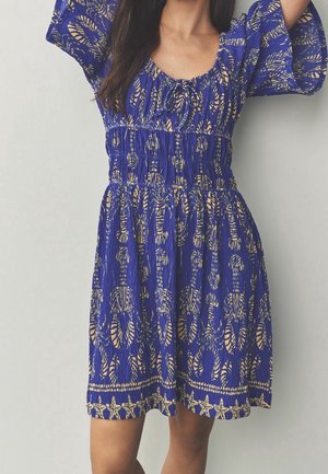 Femme portant une robe bleue longueur genou avec des motifs abstraits beiges et étoiles de mer, taille froncée et manches mi-longues, debout devant un fond uni.