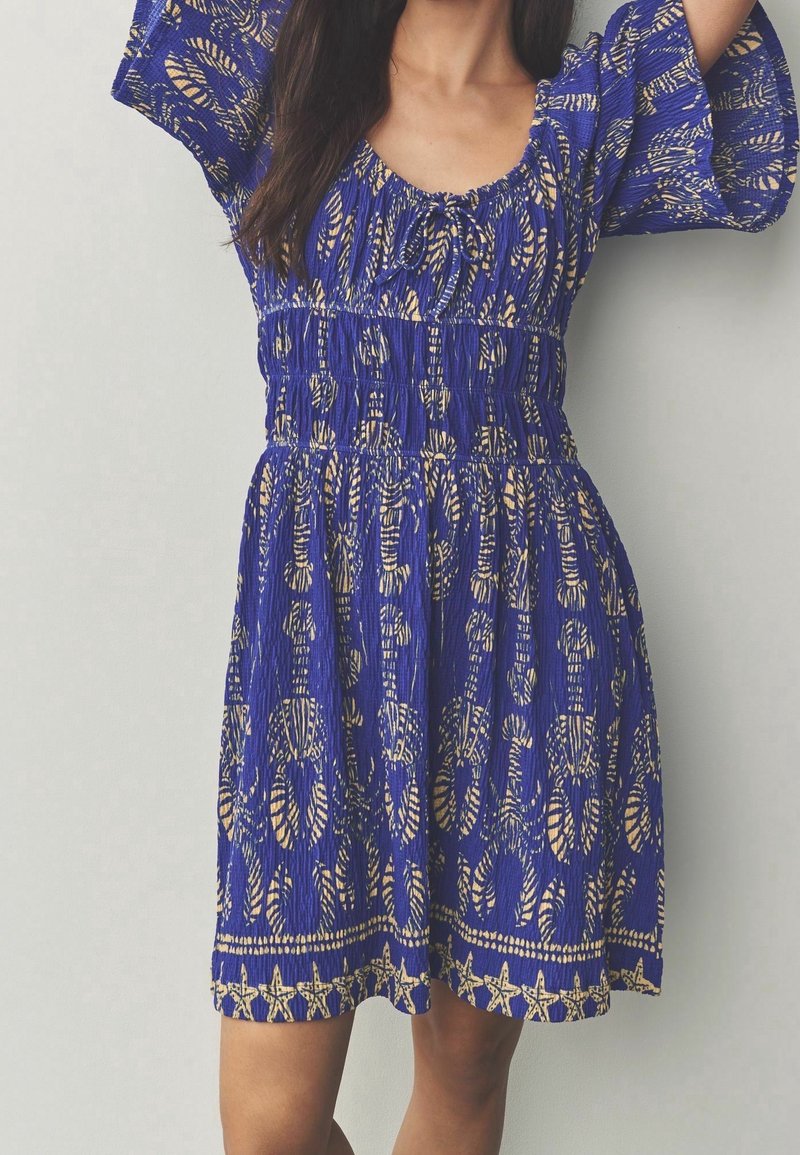 Femme portant une robe bleue longueur genou avec des motifs abstraits beiges et étoiles de mer, taille froncée et manches mi-longues, debout devant un fond uni.
