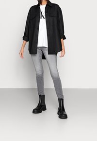 Black button-up shirt with two chest pockets, white graphic t-shirt underneath, grey skinny jeans, and black ankle boots with chunky soles.