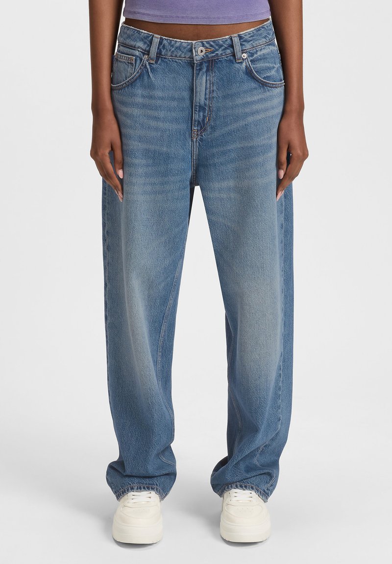 Une personne portant un jean bleu ample et des baskets blanches, se tenant droit avec les mains détendues sur les côtés.
