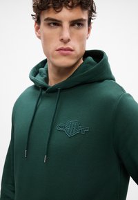 Felpa con cappuccio verde scuro realizzata in tessuto morbido, dotata di tasca a marsupio, coulisse regolabile e logo ricamato sul petto.