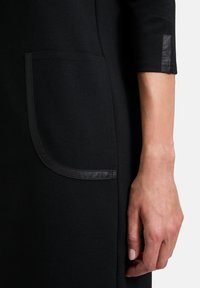 Svart klädestyg med en slät yta, som har en sidficka kantad i blank svart trim, och en åtsittande ärm med en läderdetalj.