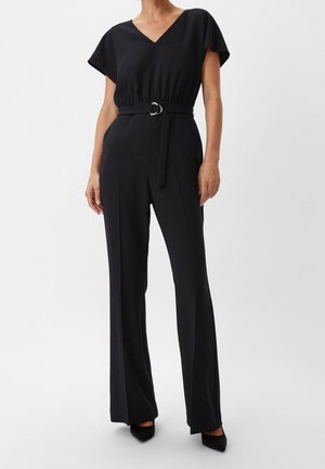 Kvinde iført en sort jumpsuit med bælte, korte ærmer, V-hals og bukser med vide ben, kombineret med sorte spidse hæle.