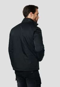 Melnā izolētā jaka ar stāvošu apkakli, rāvējslēdzēja aizdari un uzšuvumu uz kreisā pleca; gluda tekstūra un pieguļošs siluets.