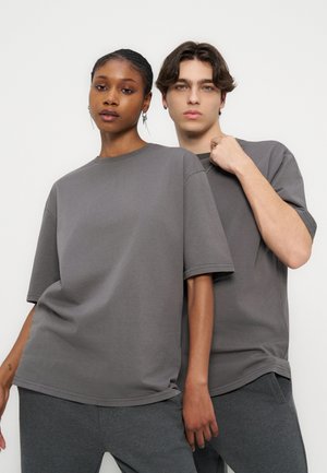 Du jauni suaugusieji dėvi atitinkančius laisvus pilkus marškinėlius ir tamsias kelnes, stovi arti vienas kito priešais paprastą šviesų foną.