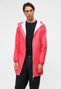 Homme portant une veste de pluie à capuche rose vif par-dessus un t-shirt noir et un pantalon noir, debout devant un fond clair uni.