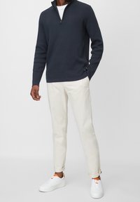 Man i en marinblå zip-sweater, vit t-shirt, krämfärgade uppvikta byxor och vita sneakers med orangea detaljer.