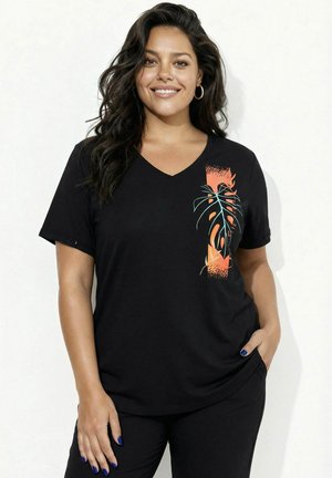 Lächelnde Frau mit langen dunklen Haaren trägt ein schwarzes V-Neck-T-Shirt mit einem orangeblauen tropischen Blattmuster, die Hände entspannt, eine in der Tasche.