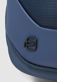 Blau strukturierte Stofftaschenecke mit einem glatten, dunkelblauen Akzent und einem schwarzen Logoemblem. Hat eine Naht entlang der Kante.