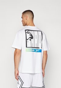 Bílý tričko s krátkým rukávem, na zadní straně s výraznou černou grafikou basketbalového míče, s akcenty zeleného a modrého loga Reebok.