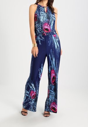 Femme portant une combinaison bleu marine sans manches à jambes larges, ornée de motifs de plumes de paon roses vives et bleues, assortie de talons noirs et d'une manchette dorée.