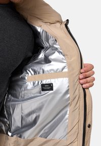 Lys beige jakke med en sølv sateng indvendig foring, med synlig merkeetikett. Yttermaterialet har en struktureret overflate og glidelås lukking.