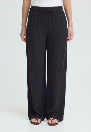 Czarne spodnie o szerokich nogawkach z elastycznym pasem i sznurkiem, noszone z białą bluzką i czarnymi sandałami, przedstawione na stojącej osobie.