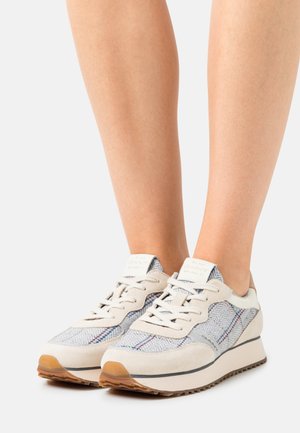 Piernas con zapatillas de deporte gris claro y beige con tela espiga y de cuadros, cordones blancos y suelas de goma sobre fondo blanco.