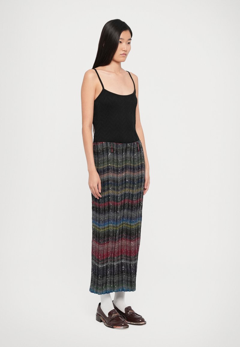 Débardeur noir à fines bretelles, associé à une jupe longue plissée multicolore avec des motifs ondulés en bleu, rouge, vert et accents argentés.