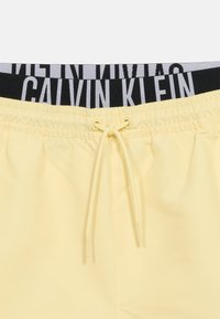 Ανοιχτό κίτρινο σορτς με ελαστική μέση και κορδόνι, πάνω από μαύρο εσώρουχο με λογότυπο Calvin Klein στη μέση.
