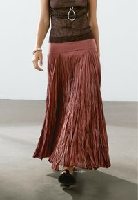 Jupe longue plissée d'un doux burgundy riche. Fabriquée en tissu léger avec un léger éclat. Taille plate. Chaussure à bout rond visible.