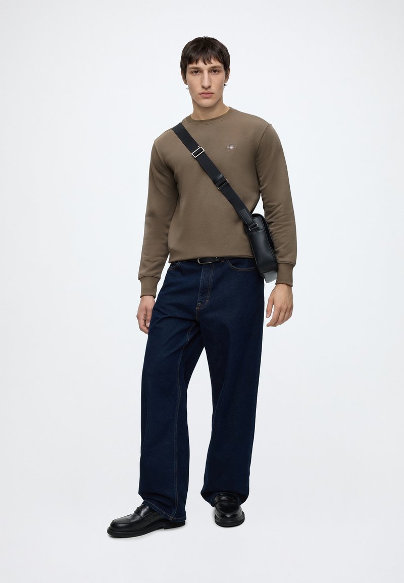 Ung mand iført olivengrøn sweatshirt, mørkeblå vidde jeans, sorte lædersko og en sort crossbody taske mod en ensfarvet lys baggrund.