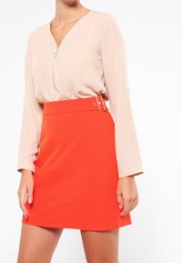 Blusa rosa chiaro con zip e maniche lunghe; abbinata a una mini gonna arancione brillante con una cintura spessa e dettagli dorati.