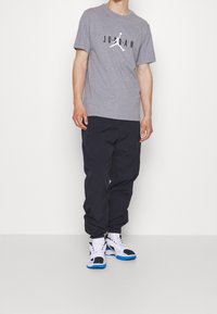 Man som bär en grå Jordan-t-shirt, svarta joggerbyxor, vita strumpor och vita sneakers med blå sulor, stående mot en helt vit bakgrund.