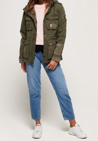 Olijfgroene utility jas met meerdere zakken, decoratieve patches en een capuchon, gecombineerd met lichtblauwe jeans met rechte pijpen en witte sneakers.