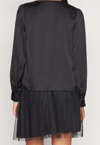 Robe noire avec une partie supérieure en satin et une jupe en tulle à volants, dotée de manches longues et de détails en bouton aux poignets.