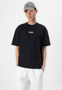 T-shirt nera in cotone a maniche corte e colletto rotondo, con un logo giallo sul petto. Abbinata a pantaloni bianchi in lino e un cappellino grigio.