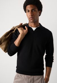 Čierny pletený sveter s výstrihom do V a rebrovanými manžetami, v kombinácii s svetlohnedým kabátom drapovaným cez rameno, modelovaný mužom s afro účesom.