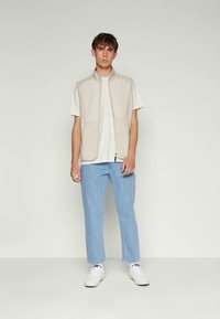 Joven de pie contra un fondo liso, vistiendo un chaleco beige con cremallera, camiseta blanca, jeans de color azul claro y zapatillas blancas.