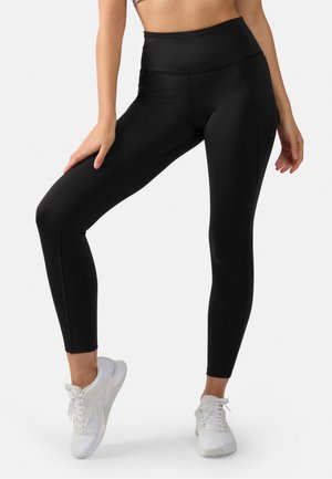 Vrouw draagt zwarte leggings met hoge taille en witte sportschoenen, poseert met één been gebogen en hand op dij rustend.