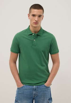 Jeune homme aux cheveux courts portant un polo vert à manches courtes et un jean bleu, debout les mains dans les poches devant un fond uni.