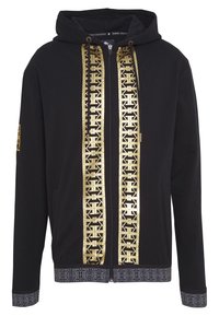 Sudadera negra con un logo dorado impreso en la parte frontal, acentos texturizados en los puños y un dobladillo con patrón. Cuenta con una cremallera y capucha con cordones.