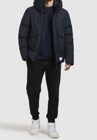 Granatowa puchowa kurtka z kapturem, z dwoma przednimi kieszeniami i zapięciem na suwak. Noszona na ciemnym swetrze i czarnych spodniach, do tego białe trampki.