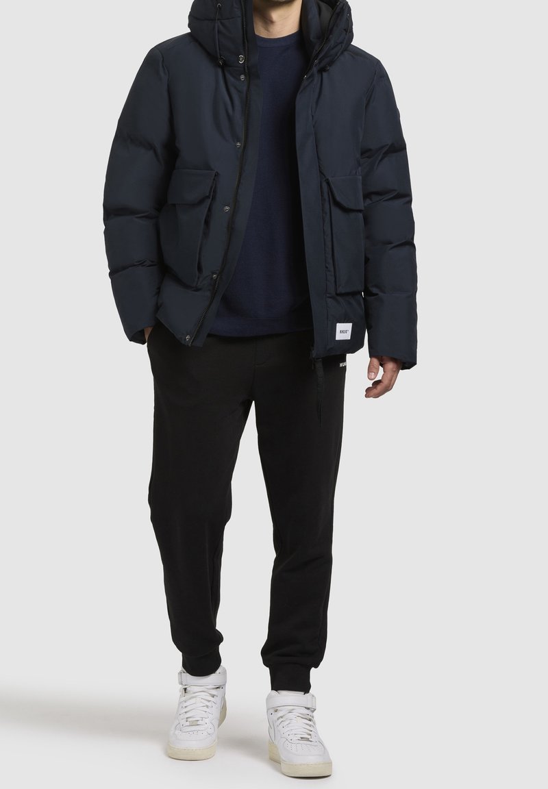 Granatowa puchowa kurtka z kapturem, z dwoma przednimi kieszeniami i zapięciem na suwak. Noszona na ciemnym swetrze i czarnych spodniach, do tego białe trampki.