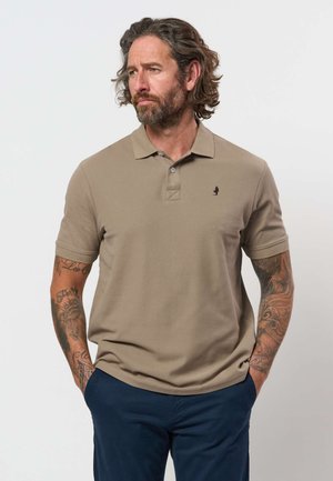Homme aux cheveux bouclés et barbe, portant un polo beige et un pantalon bleu marine avec des tatouages sur les deux bras, mains dans les poches, regardant vers la gauche.