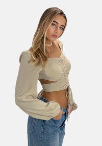 Blusa beige de manga larga con un frente fruncido y diseño recortado, combinada con jeans de mezclilla azul de cintura alta. Textura suave, ajuste relajado.