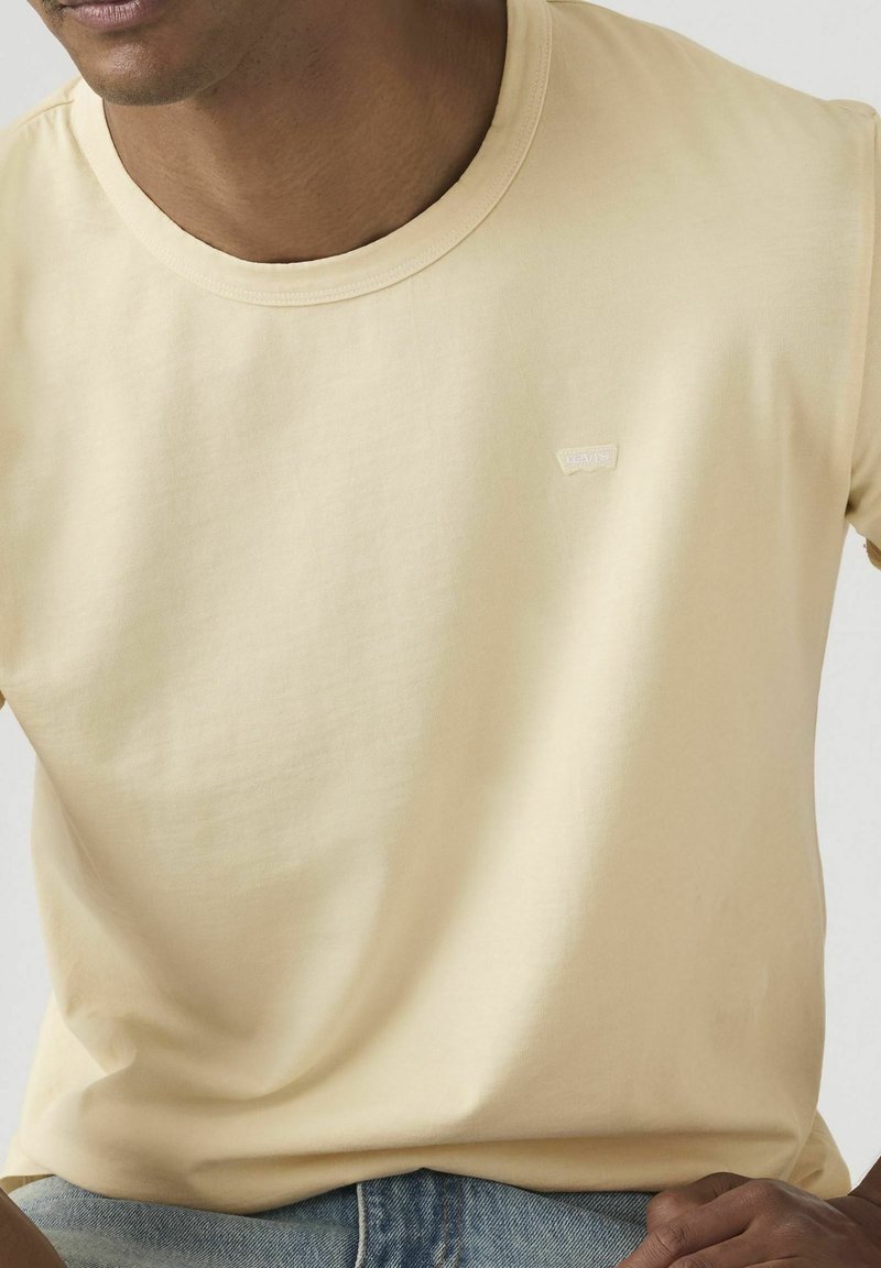 Uomo che indossa una maglietta gialla chiaro a girocollo senza decorazioni e jeans blu, con un piccolo logo rettangolare sul petto a sinistra.