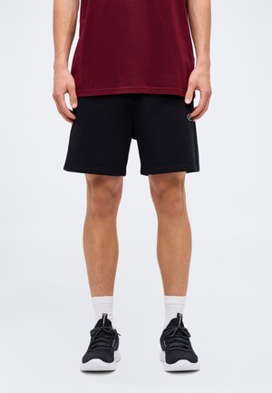 Man wearing maroon shirt, black shorts, white socks, and black athletic shoes standing against a plain light background.