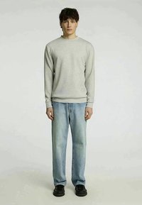 Suéter cinza claro com punhos canelados, combinado com jeans azuis desbotados. O modelo está em sapatos pretos contra um fundo simples.