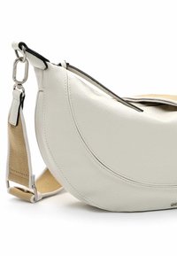 Bílá kožená crossbody taška s zakřiveným designem, kontrastním černým lemováním a béžovým textilním popruhem. Disponuje zipovým uzávěrem a hladkou texturou.