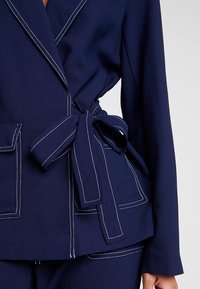 Námořnická modrá zavinovací bunda vyrobená z měkké látky, se bílým kontrastním prošitím, předními kapsami a zavazováním v pase.