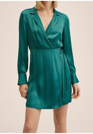 Femme portant une robe portefeuille à manches longues de couleur sarcelle, avec col et ceinture sur le côté, debout devant un fond clair uni.