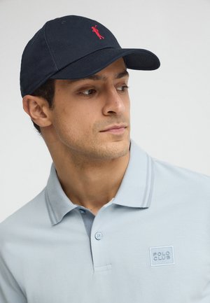 Homme portant un polo bleu clair avec le logo "Polo Club" et une casquette bleu marine avec un logo de joueur de polo brodé en rouge, regardant sur le côté.