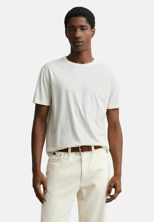 Jonge man die een eenvoudige witte kortemouwen T-shirt draagt, ingestopt in gebroken witte spijkerbroek met een bruine riem, staand tegen een witte achtergrond.