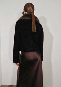 Černá kordurová bunda s kožešinovým límcem, zkrácený střih, zkombinovaná s hladkou hnědou saténovou sukní. Vlasy stažené do ohonu. Minimalistické pozadí.