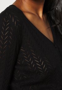 Gros plan d'une personne portant un cardigan en maille noire avec un motif en chevrons et des boutons, montrant la zone du cou et des épaules.