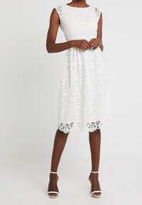 Robe en dentelle blanche avec motifs floraux, longueur genou, col rond, manches courtes volantées et ourlet festonné. Associée à des talons blancs.