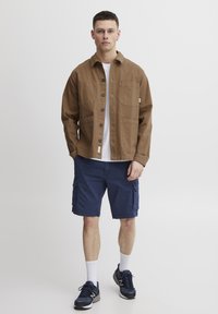 Veste utilitaire en coton marron avec des fermetures à bouton et des poches poitrine, associée à un short cargo bleu marine et des chaussures de sport noires.